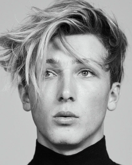 Headshot of a young adult with light hair and a black turtleneck against a gray background, showcasing a modern, minimalist portrait.