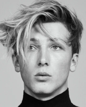 Headshot of a young adult with light hair and a black turtleneck against a gray background, showcasing a modern, minimalist portrait.