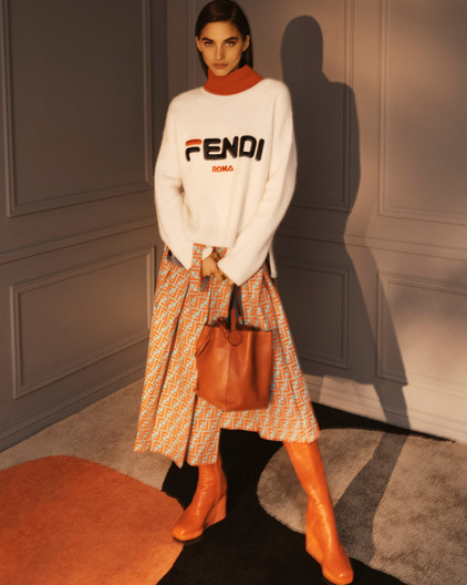 Fashion shoot of a woman in a Fendi sweater and patterned skirt holding a leather bag, standing indoors with soft, diffused lighting.
