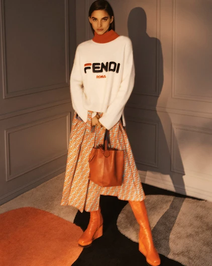 Fashion shoot of a woman in a Fendi sweater and patterned skirt holding a leather bag, standing indoors with soft, diffused lighting.
