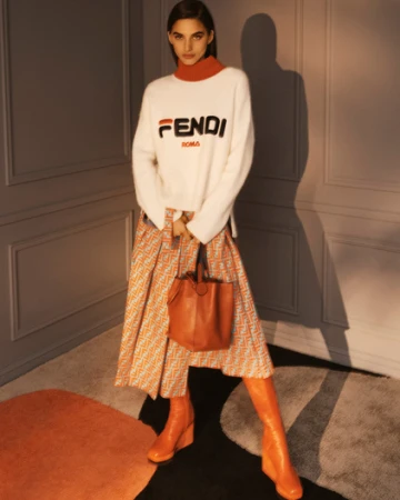 Fashion shoot of a woman in a Fendi sweater and patterned skirt holding a leather bag, standing indoors with soft, diffused lighting.