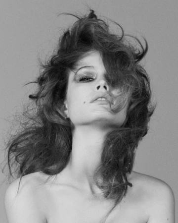Monochrome portrait of a woman with tousled voluminous hair looking downward with dramatic eye makeup against a grey background.