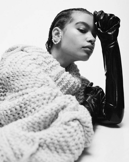 Monochrome portrait of a young adult with braided hair wearing a knit sweater and black gloves against a bright white background.
