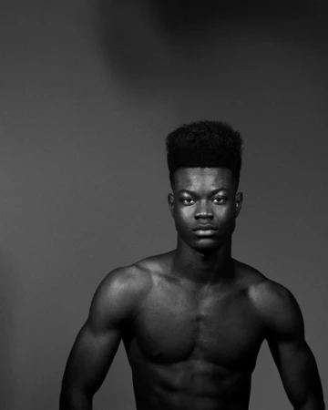 Monochrome portrait of a muscular, shirtless Black man with a high-top fade hairstyle against a grey backdrop in a photo studio.