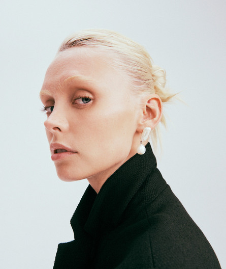 Close-up portrait of a fair-skinned woman with blonde hair wearing a black jacket and pearl earrings against a white background.