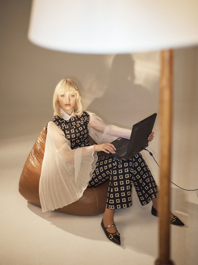 Woman in patterned outfit sits in a beanbag chair and uses a laptop in an indoor setting with a lamp.