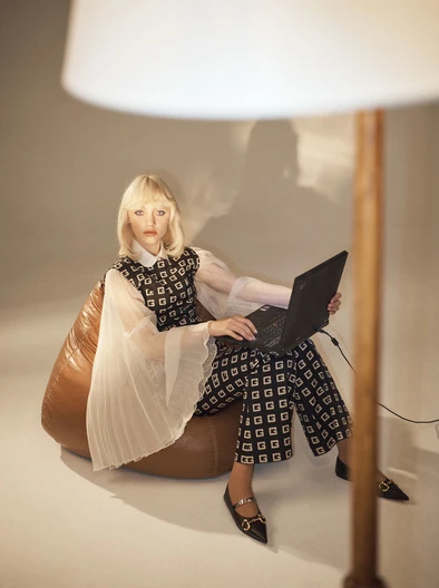 Woman in patterned outfit sits in a beanbag chair and uses a laptop in an indoor setting with a lamp.