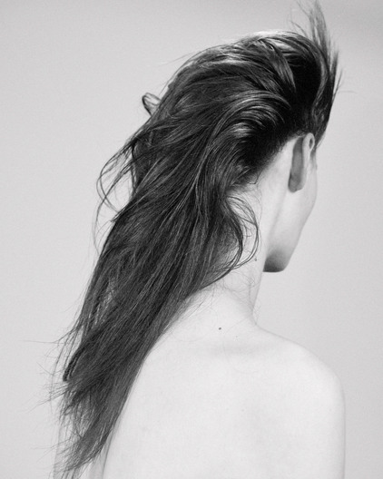 Back view of a fair-skinned adult female with long, dark hair styled upwards in a beauty fashion portrait against a solid grey background.