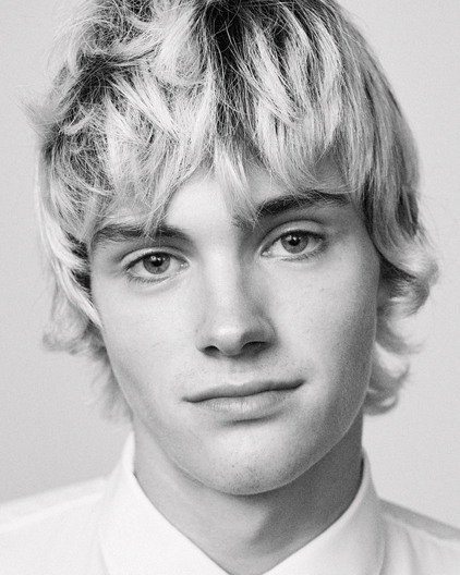 Striking black and white portrait of a young adult male with blond wavy hair wearing a white collared shirt against a neutral background.