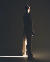 A person standing with back turned, wearing a dark jacket and trousers, illuminated by a beam of light in a dark studio.