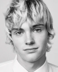 Close-up portrait of a young man with tousled blonde hair wearing a white collared shirt against a plain background.