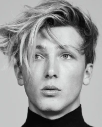 Close-up monochrome portrait of a young man with messy styled hair wearing a dark turtleneck against a plain grey backdrop.