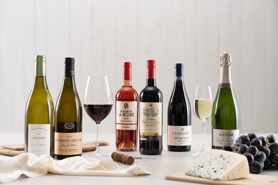 Wine assortment features white, rose, and red bottles paired with cheese, grapes, and a corkscrew on a white table against a bright backdrop.