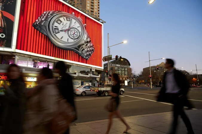 Large billboard displays wristwatch over busy city street with blurred pedestrians and cars; dusk sky in background.