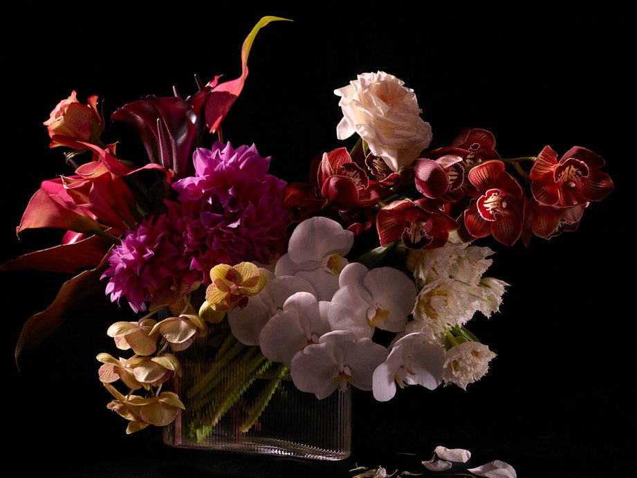 Still life photograph of a vibrant floral arrangement featuring dahlias, orchids, lilies, and roses in a clear glass vase against a black background.