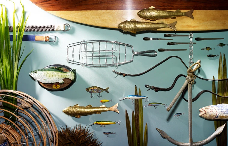 Arrangement of fishing-themed objects on a wall: fishing lures, fish mounts, tackle, and a rustic anchor against a blue background.