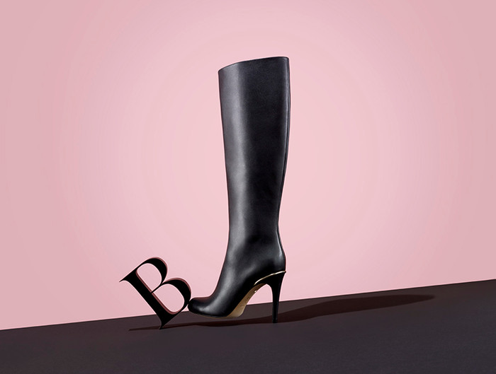 Black leather knee-high heeled boot stands on a black surface with a large letter 'B' against a split pink backdrop.