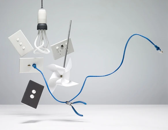 Still life of floating electrical objects in a studio setup showcasing plugs, sockets, a lightbulb, a cable, pliers, and a pinwheel.