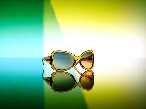 Yellow sunglasses with gray gradient lenses are reflected on a glossy surface with green and yellow background lighting. Studio shot.