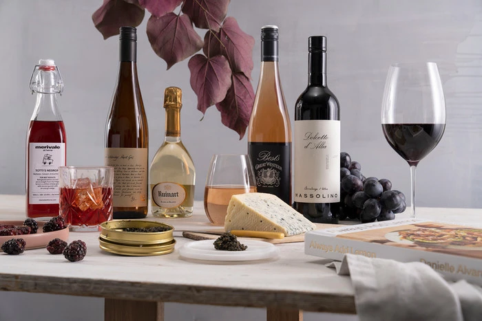 A still-life arrangement features wine bottles, wine glasses, blackberries, caviar, cheese, and a cookbook on a white wooden table.