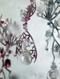 Close-up of ornate coral-inspired earrings with rubies, diamonds, and pearls hanging behind a water-streaked glass surface.