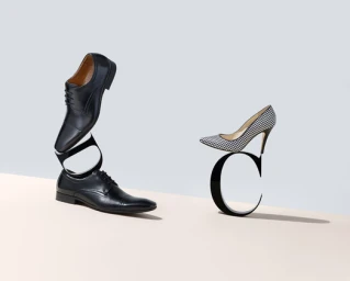 Black leather dress shoes and a houndstooth stiletto pump balanced on sculptural letters against a light studio background.