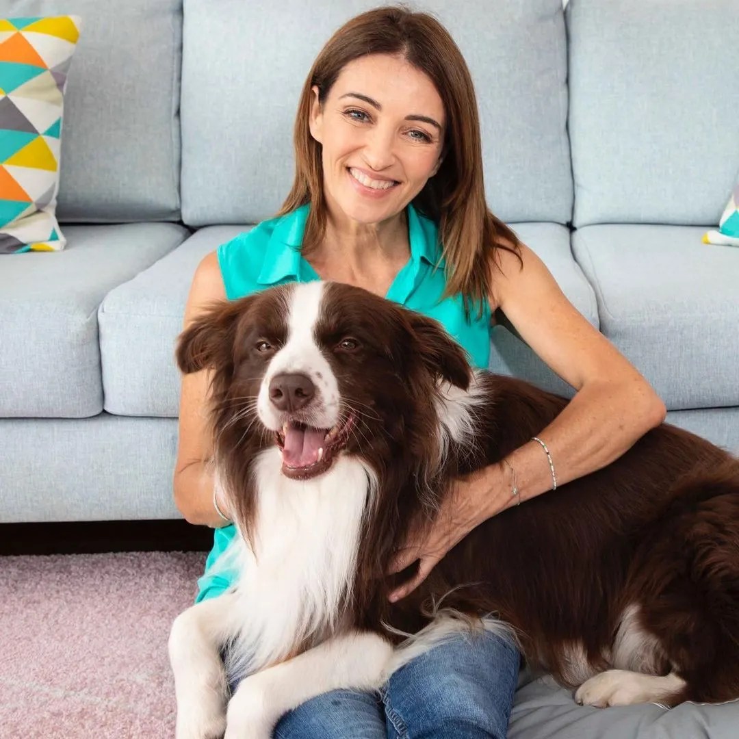 Adult woman smiles while seated with her pet dog in a modern indoor setting with a light blue couch and light purple carpeting.