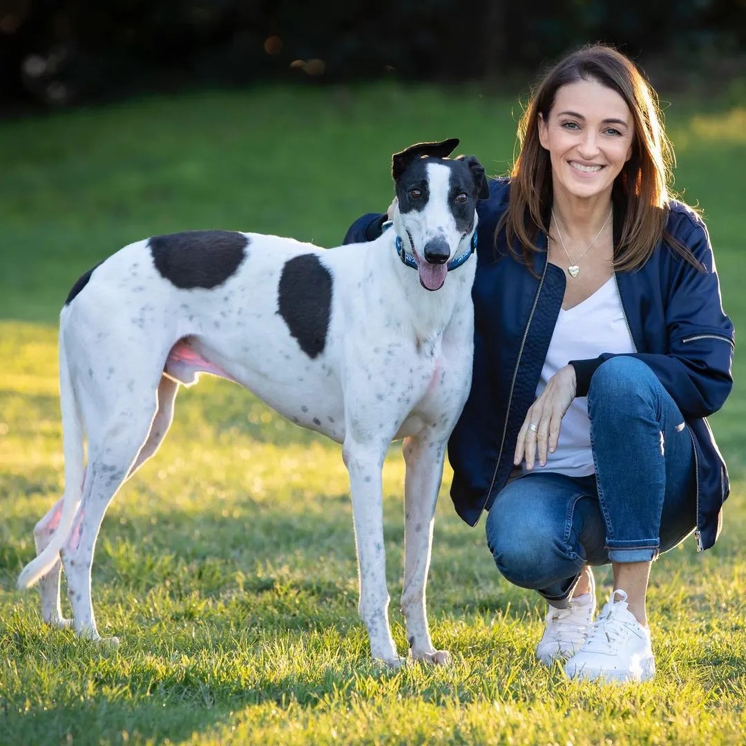 Woman crouches next to a white and black spotted greyhound dog in a grassy outdoor setting, both looking toward the camera.