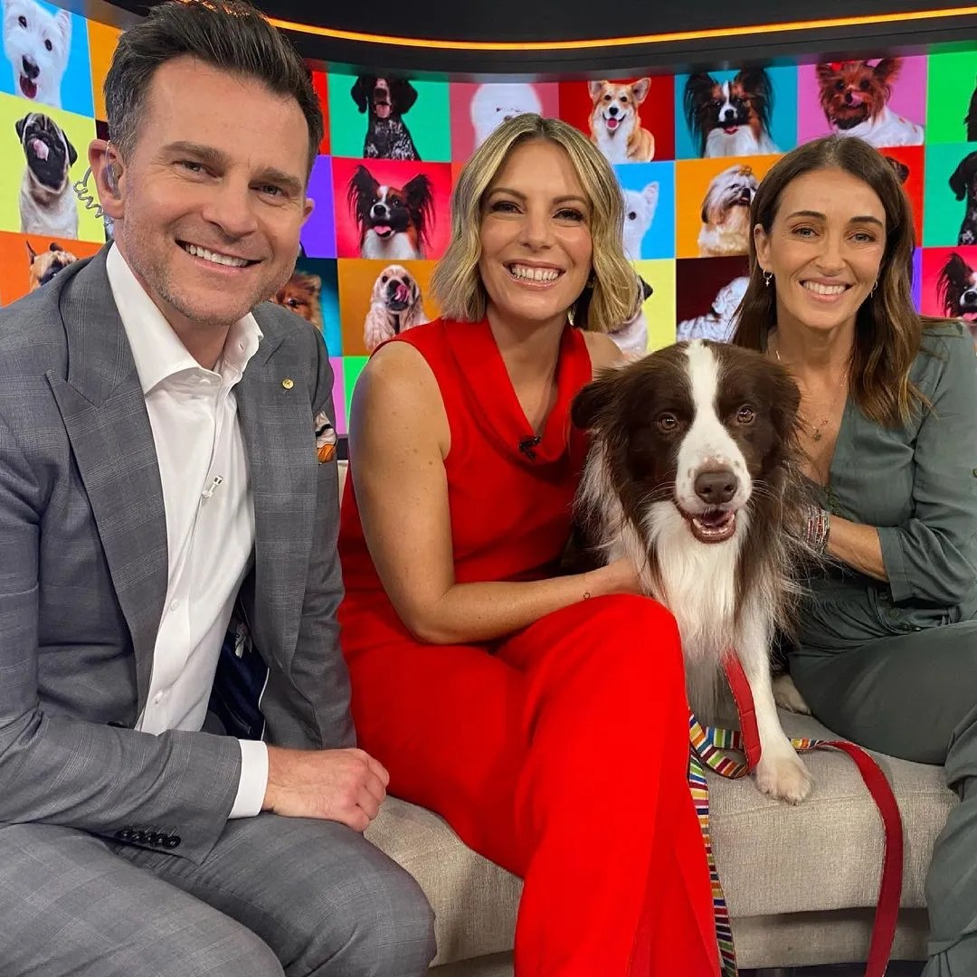 Three smiling people with a dog sit indoors against a colorful backdrop; the central woman is hugging the medium-sized dog.