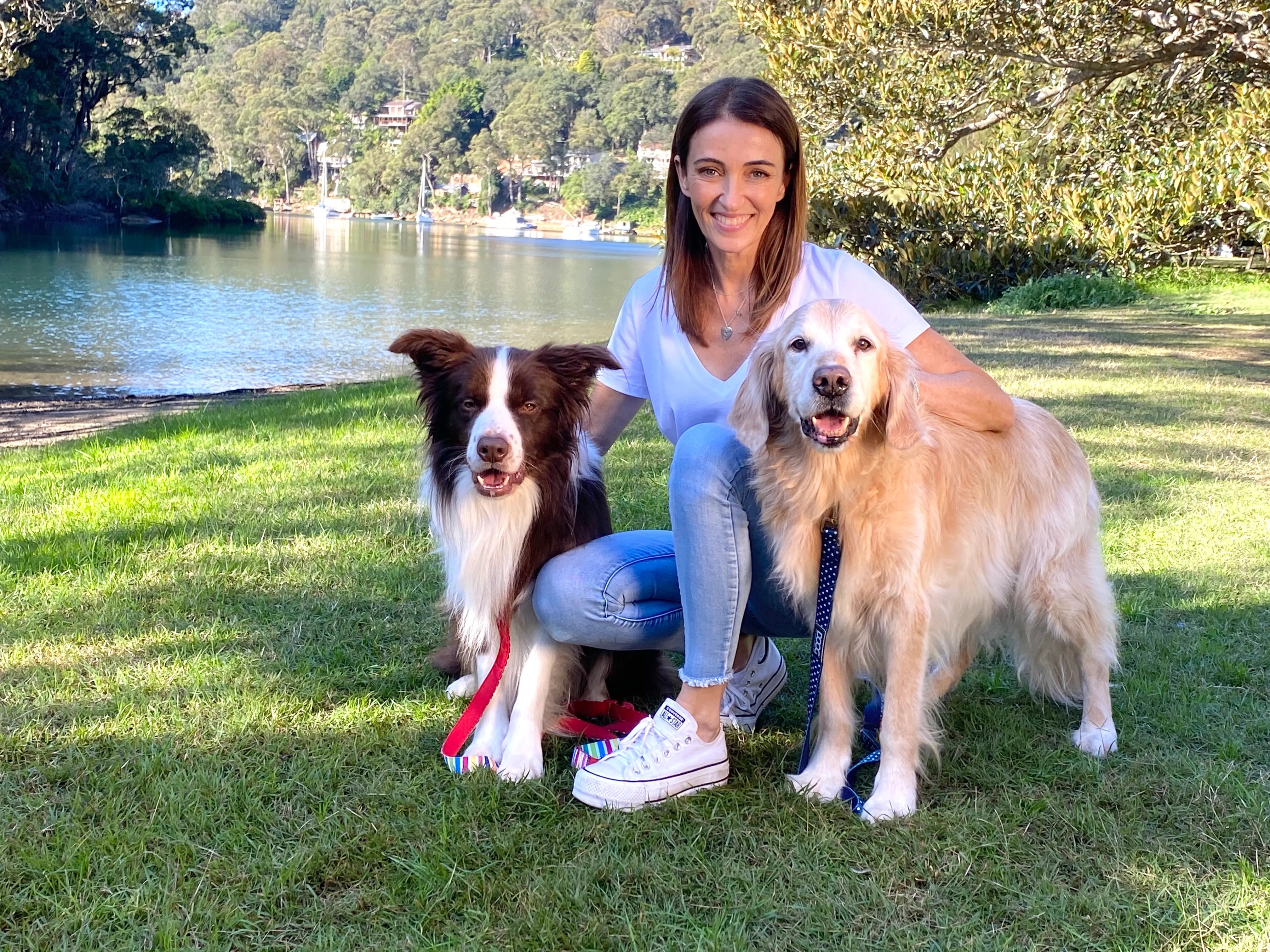 A woman smiles while crouching with two dogs on grass near a lake with wooded hills in the background on a sunny day.