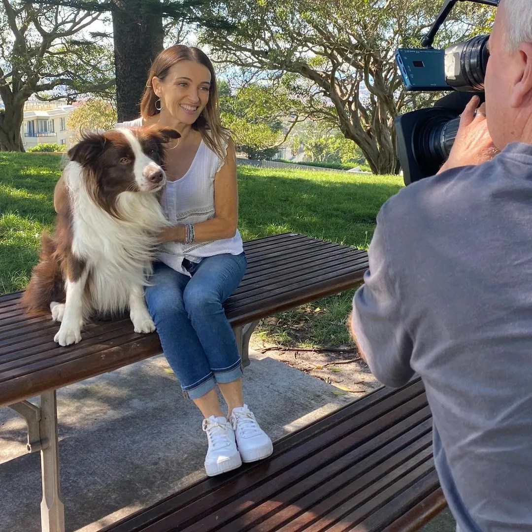 Woman and dog sit on a park bench being filmed by a cameraman in a park with green grass and trees on a sunny day.