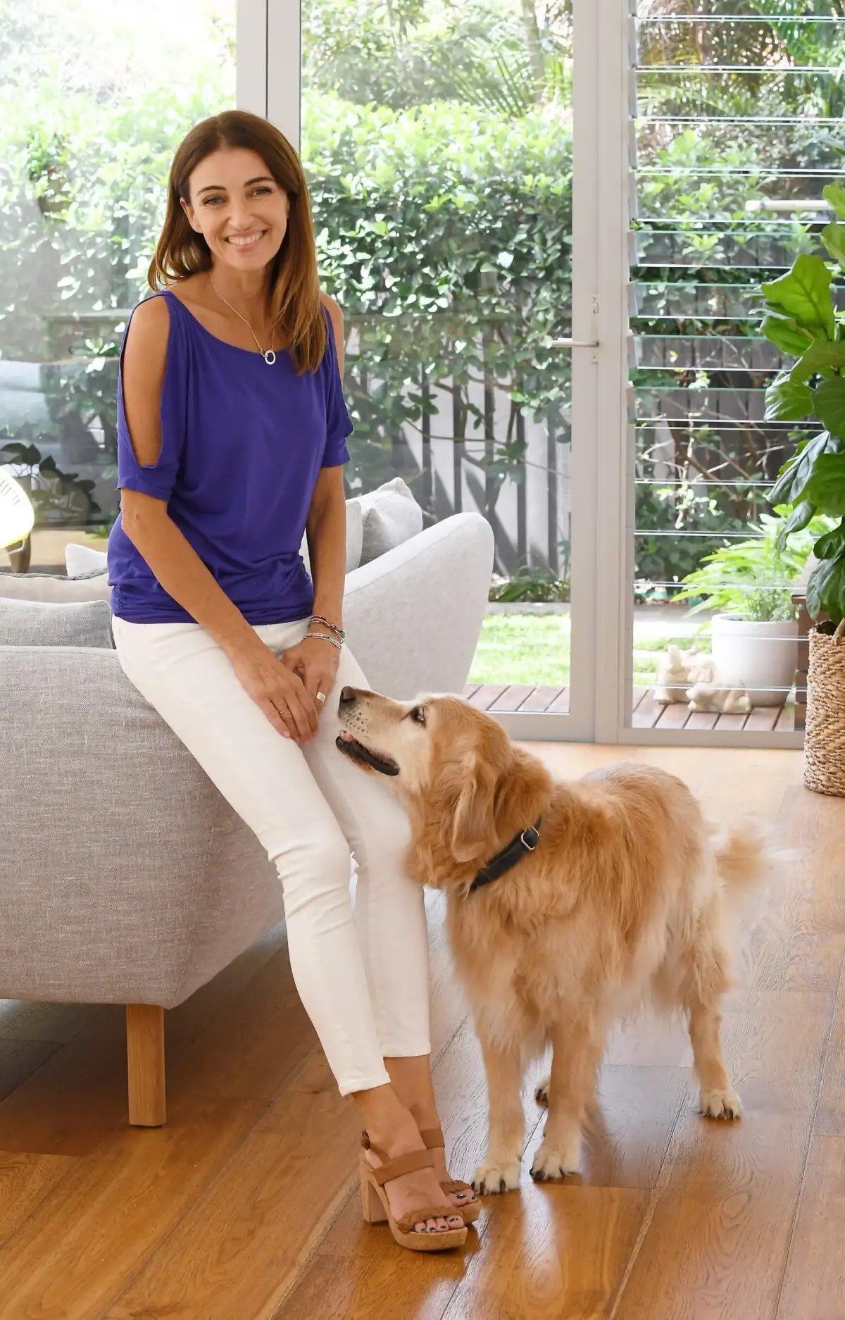 Woman in blue top poses with Golden Retriever dog indoors by a window with green foliage background. Light wood flooring.