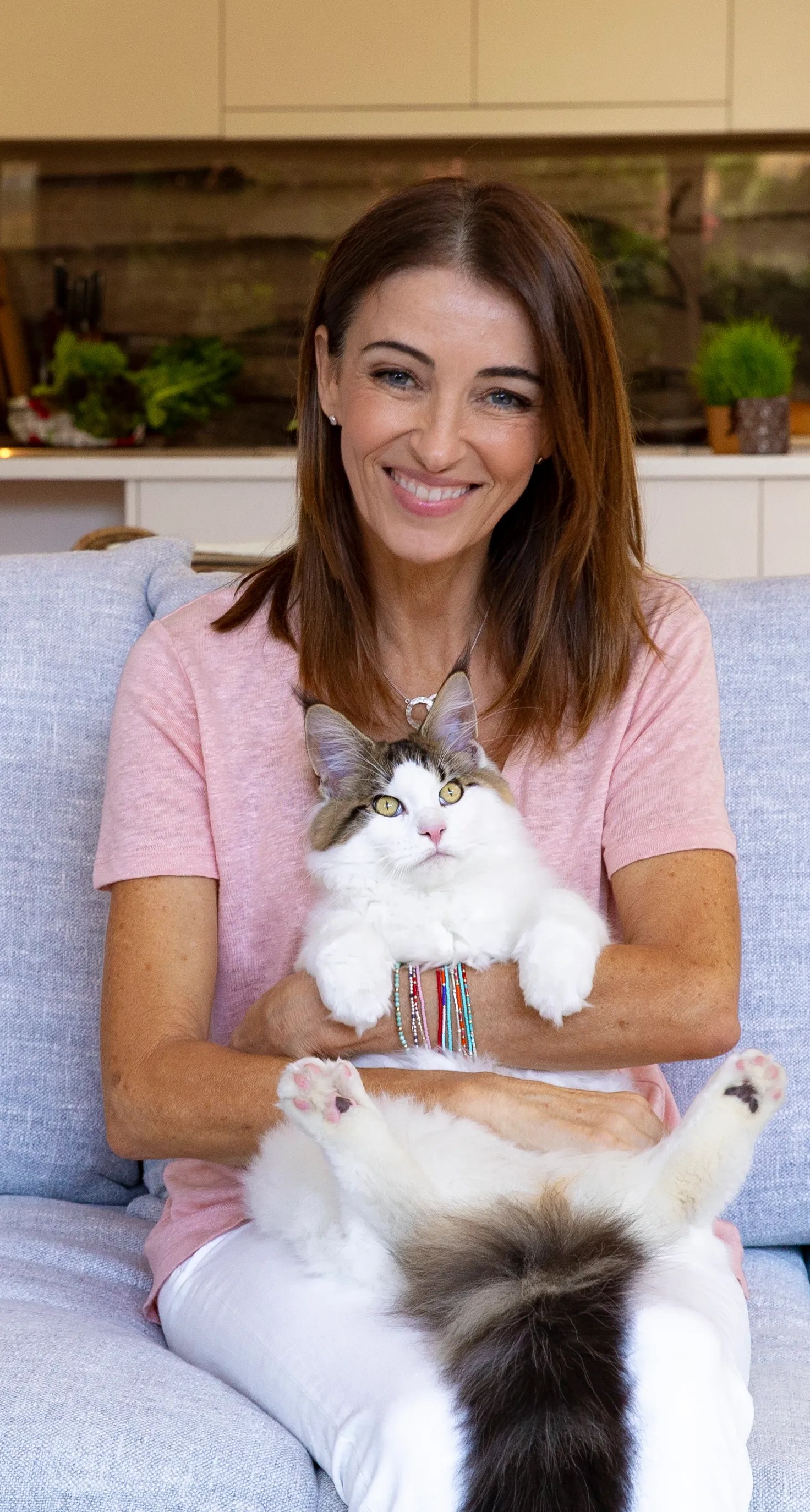 Adult woman smiles while sitting on a couch and holding a large white cat with brown markings indoors in front of a kitchen area.