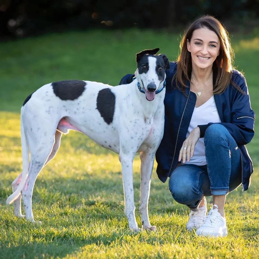 Dr Katrina Warren crouching in a grassy park next to a tall white and black spotted greyhound dog outdoors.