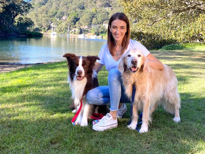Dr Katrina Warren kneeling on a grassy waterfront park in Sydney with two dogs by her side.