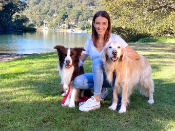 Dr Katrina Warren kneeling on a grassy waterfront park in Sydney with two dogs by her side.