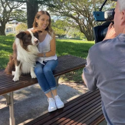 Dr Katrina Warren seated on a park bench next to a border collie while being filmed by a camera operator in Sydney.