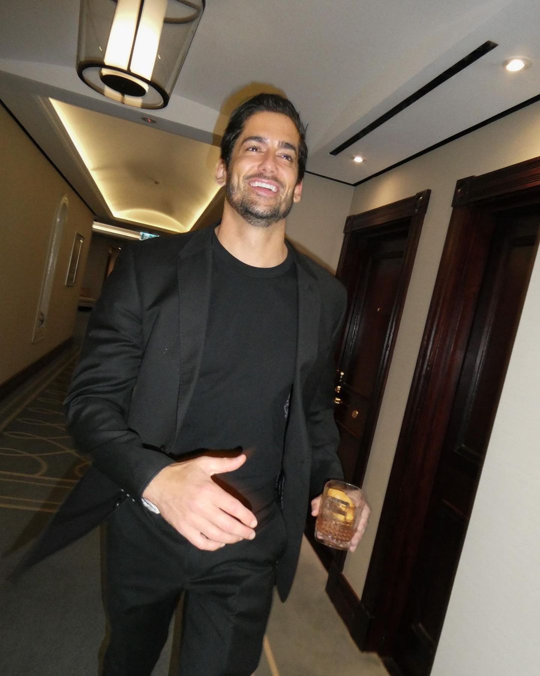 Smiling man in a black suit holds a glass with ice and lemon in a hallway with doors and ceiling lights. Dark and modern look.