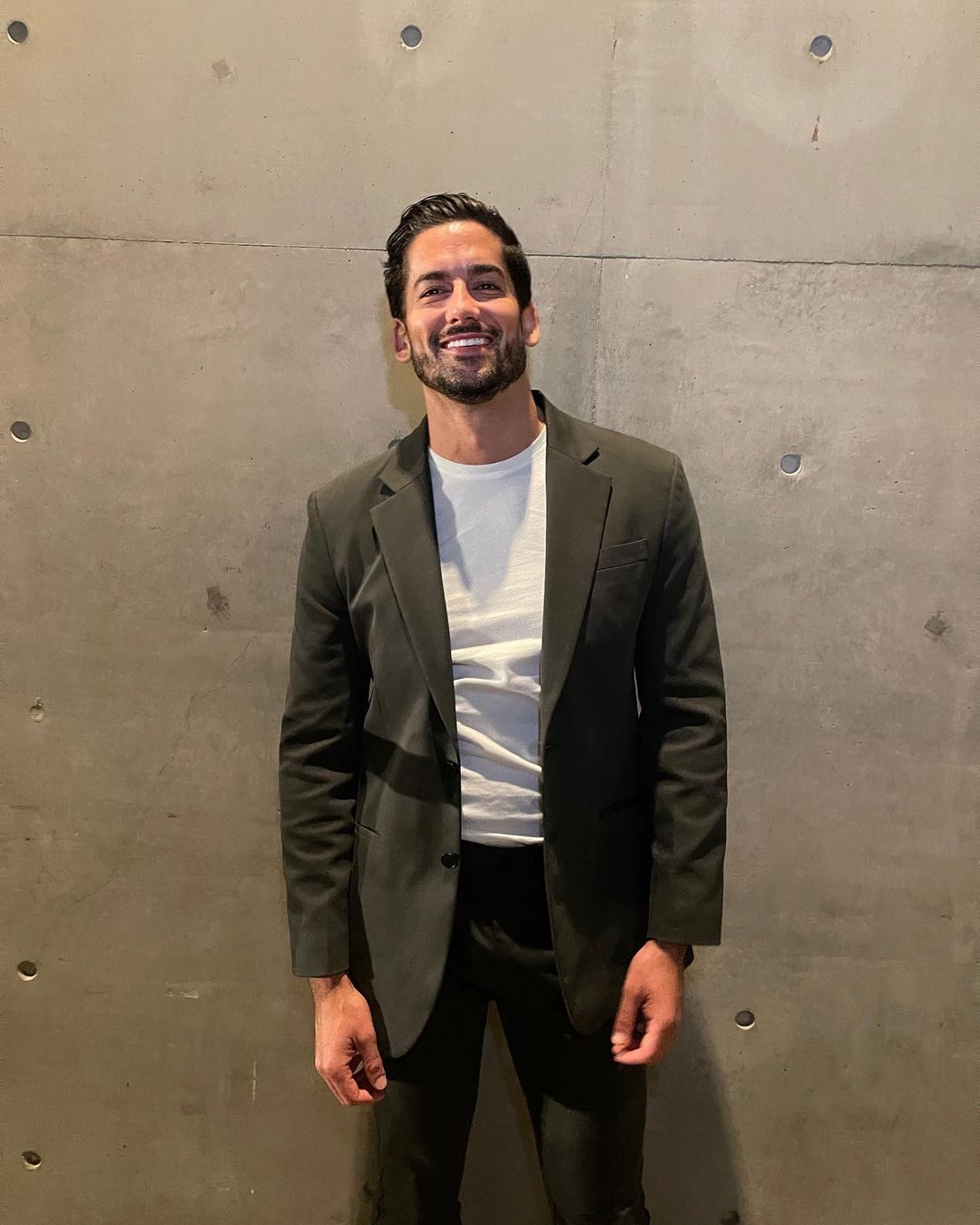 Portrait of a smiling adult male wearing a dark suit and a white t-shirt, standing indoors in front of a concrete wall.