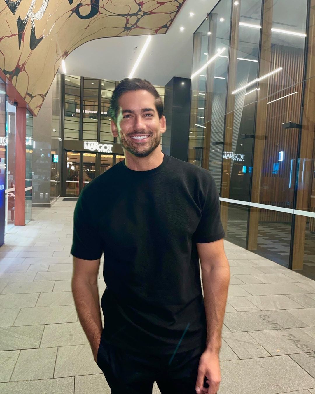 A smiling adult male in a black t-shirt stands indoors with a modern urban backdrop of glass windows and a decorative ceiling.
