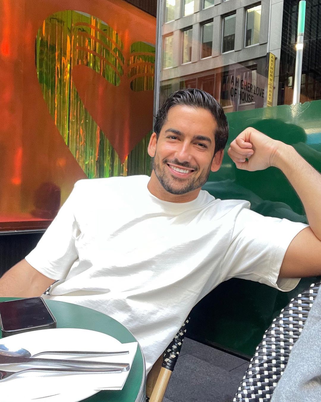 Smiling adult male in a white t-shirt is sitting outside at a green table with a drink in daylight with an urban background.