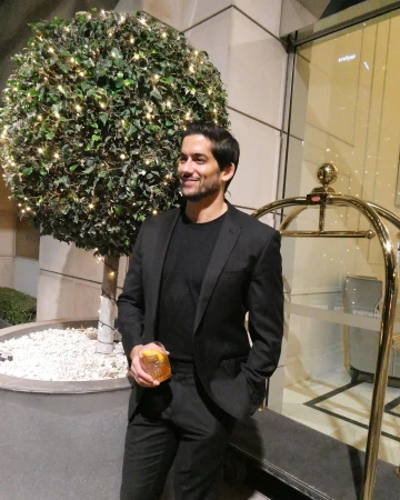 Handsome man in a black suit holds a cocktail next to a plant with fairy lights in an upscale outdoor setting at twilight.