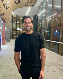 Man wearing a black t-shirt smiling outdoors on a paved street at night in front of modern glass buildings