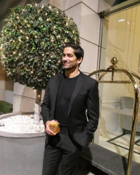 Man in black suit holding a drink outside hotel lobby with topiary tree and brass luggage cart