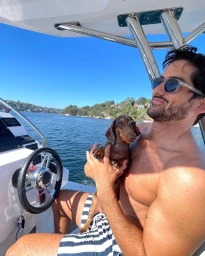 Man wearing sunglasses and striped swim trunks holding a small dachshund dog on a boat in Sydney