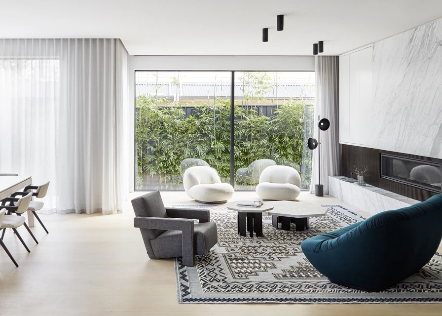 Contemporary living room interior with modern armchairs, hexagonal coffee tables, patterned area rug, and a wall of glass overlooking greenery.