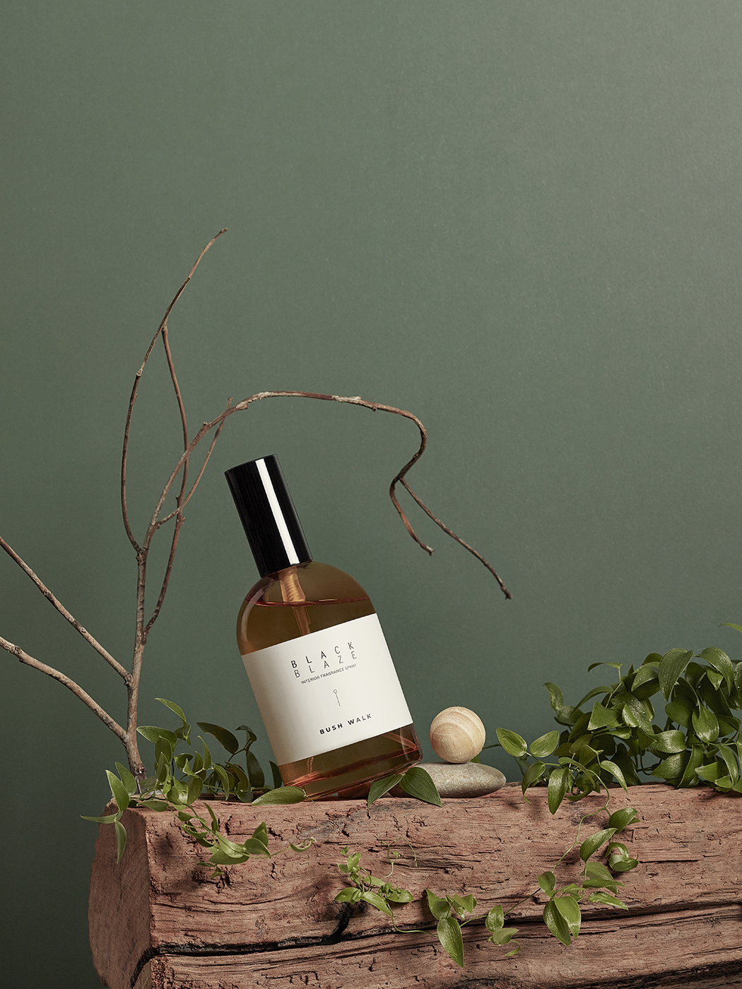 A perfume bottle sits on a wooden log alongside greenery and a smooth stone against a muted green background, evoking nature and calm.