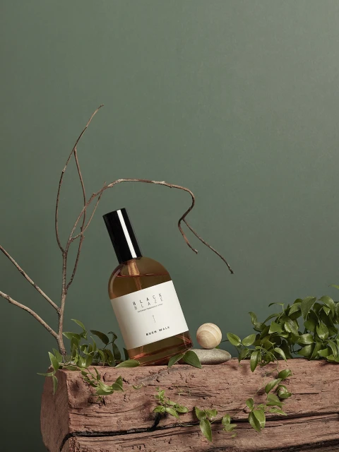 A perfume bottle sits on a wooden log alongside greenery and a smooth stone against a muted green background, evoking nature and calm.
