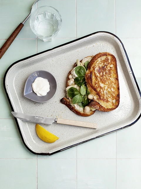 Overhead shot of a sandwich with greens, a bowl of white sauce, knife, lemon, and a water glass on a white tray against a blue tile background.