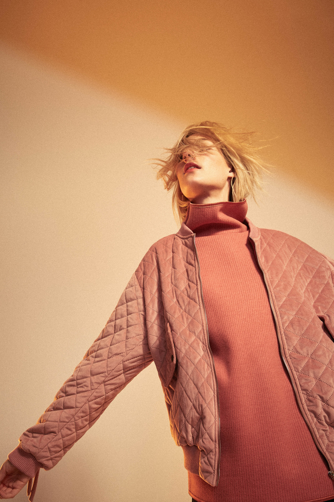 Blond woman with windblown hair poses in a pink quilted jacket and turtleneck against a peach background in a fashion portrait.
